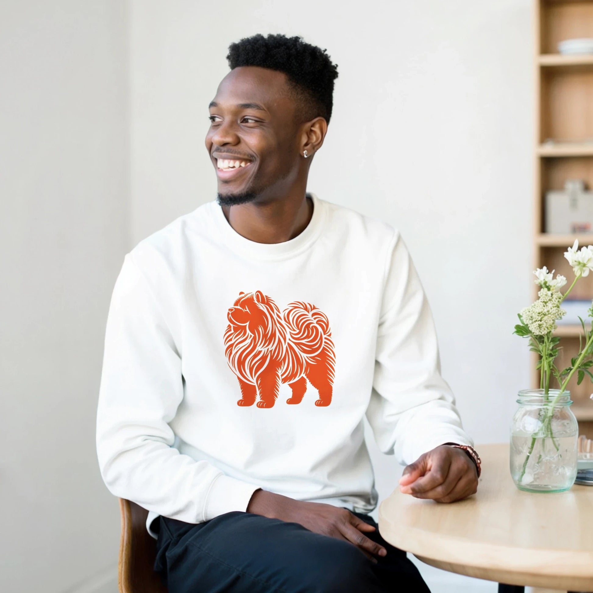 Man wearing a white sweatshirt with an orange chow chow dog design, sitting in a room with a table and flowers.