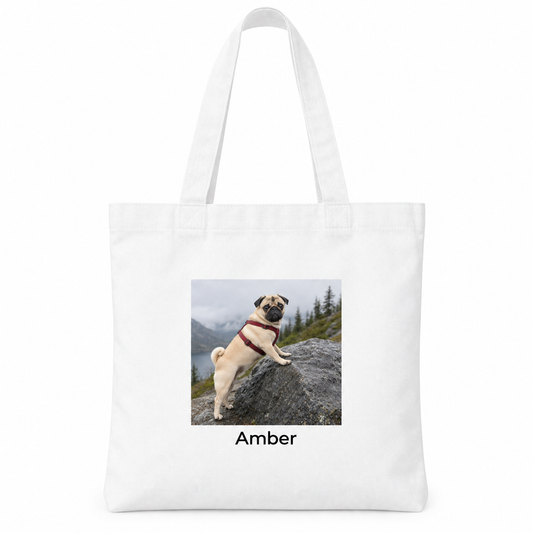 A white canvas tote bag with long handles, featuring a printed square photo of a pug dog on a rock outdoors with the name 'Amber' printed below the photo.